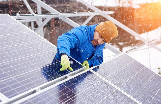 Guy with the help of tools installs solar panels in snowy weather. He is dressed in uniform and gloves