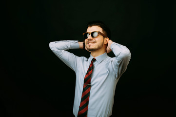 Fototapeta premium Cheerful happy young businessman smiling and and keeping his hands behind his head. Young successful man in casual wearing sunglasses over dark background.