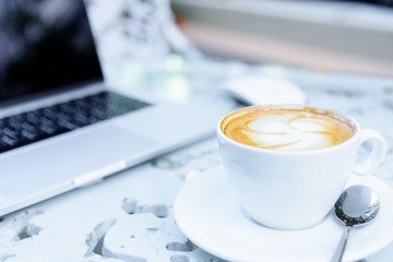 A white cup of coffee and laptop ready to working in the morning with relaxing and yellow lighting bokeh background.