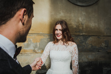 Wedding couple is walking in the old city. Stone walls of ancient town on background. Bride with hair  down is looking at groom in grey suit and bow tie. Lace elegant dress. Romantic love sight.