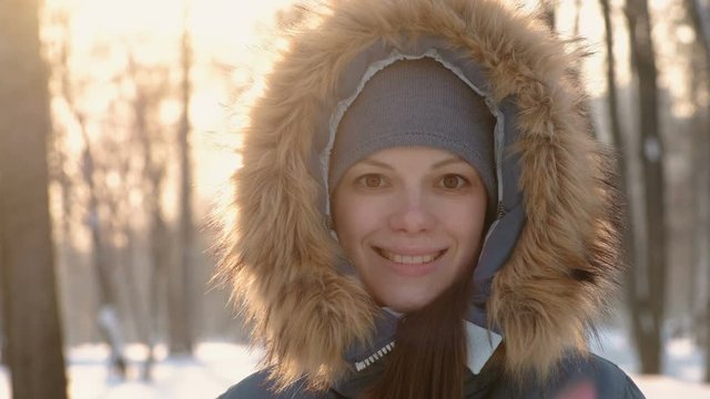Close-up woman face in fur hood looking in camera and smiling in winter sunny park.