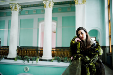 Brunette girl in green fur coat in old hall with column and railings.