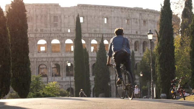 Three young friends tourists riding bikes in colle oppio park in front of colosseum on road with trees at sunset in Rome slow motion
