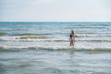 Fototapeta na wymiar Woman bruenette with long hair in blue bikini walking in the sea water with waves. Side view.