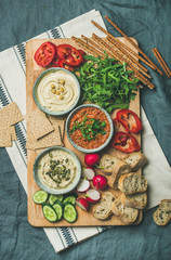 Vegan snack board. Various Vegetarian dips hummus, babaganush and muhammara with crackers, bread and fresh vegetables on wooden board over grey linen background. Clean eating, dieting food concept