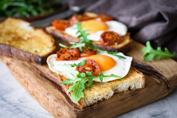 Healthy breakfast toast with egg, roasted tomato and arugula on rustic wooden cutting board....