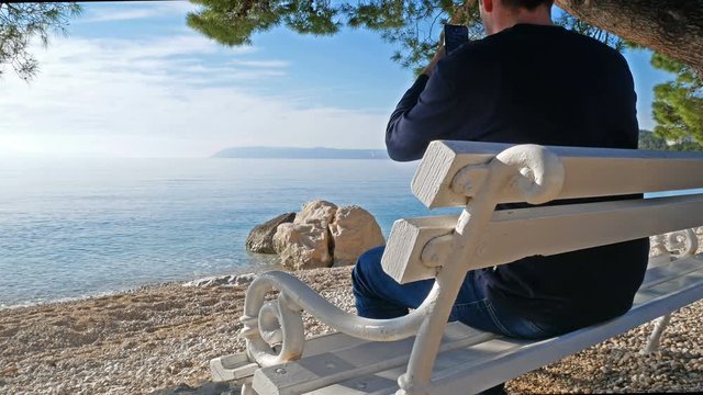 Young caucasian man sitting on wooden bench under pine tree and taking picture with mobile phone of sea landscape