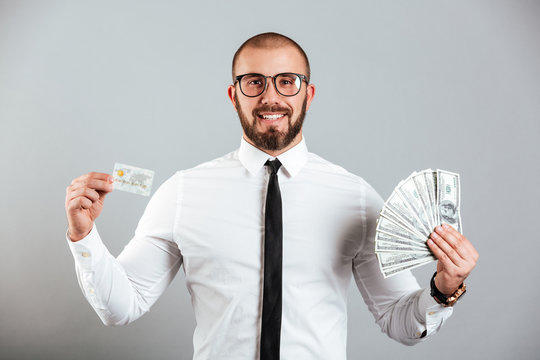 Photo of rich man 30s in glasses and tie showing credit card and fan of dollar bills, isolated over gray background