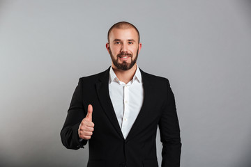 Portrait of pleased masculine man in business suit posing on camera with thumb up, isolated over gray background