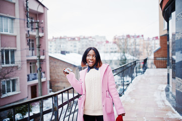 African american girl in red hat and pink coat at street of city against building on winter day with mobile phone.