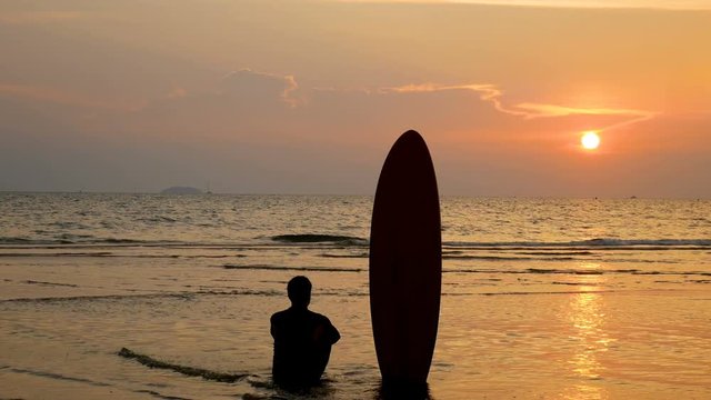 4K. silhouette of surfer man sitting on the sea beach with long surf boards at sunset on tropical beach