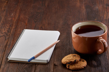 Cup of tea with cookies, workbook and a pencil on a wooden background, top view