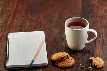 Cup of tea with cookies, workbook and a pencil on a wooden background, top view