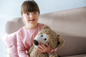Smiling child with down syndrome sitting on sofa and holding teddy bear