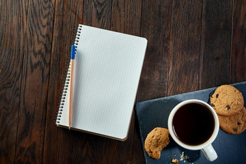 Cup of tea with cookies, workbook and a pencil on a wooden background, top view