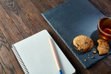 Cup of tea with cookies, workbook and a pencil on a wooden background, top view