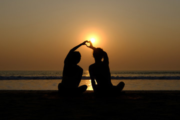 Couple's silhouette drawing heart with their hands on the beach at sunset