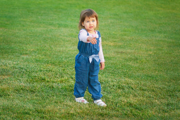 A little girl with blond hair 3 years old is standing on a green meadow and is pointing at something, copyspase