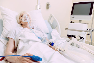 Difficult breathing. Tired elderly lady wearing an oxygen mask lying in a hospital bed with a heart rate medicine equipment.