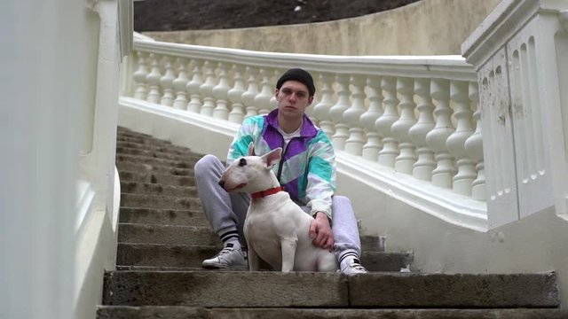 Portrait of serious male animal owner in sportswear petting his purebred bull terrier, while sitting on stairway outdoors slow motion