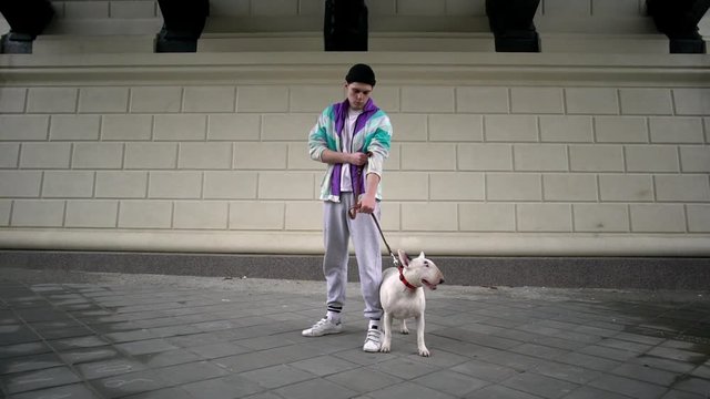 Portrait of dangerous man in sporty clothing holding his purebred bullterrier dog on leash, while standing under bridge in urban area slow motion