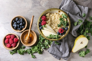Vegan quinoa porridge with kale, strawberries, blueberries, sliced pear, honey on dipper in bowl with ingredients above and cloth napkin over grey texture background. Top view, space. Healthy eating