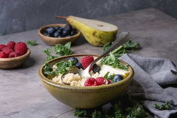 Vegan quinoa porridge with kale, strawberries, blueberries, sliced pear, honey on dipper in ceramic bowl with ingredients above and cloth napkin over grey kitchen table. Healthy breakfast