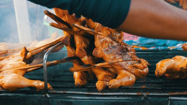 Whole chicken carcass grill strung on wooden stick grilling on the grill. Street Food Thailand. Men's hands turn over the grilled chicken