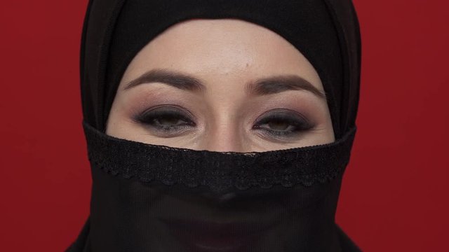 Macro portrait of middle eastern woman with smoky eyes visage in black niqab smiling on camera, over red background. Concept of emotions
