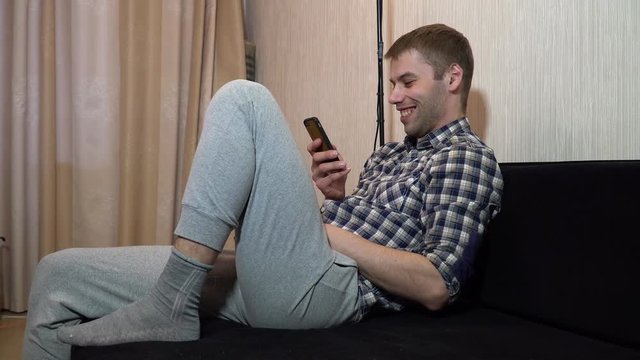 man is sitting on the couch looking into the smartphone and laughing
