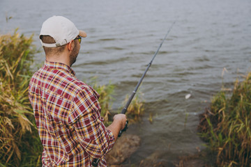 Back view Young unshaven man with a fishing rod in checkered shirt, cap, sunglasses casts bait and fishing on lake from shore near shrubs and reeds. Lifestyle, recreation, fisherman leisure concept