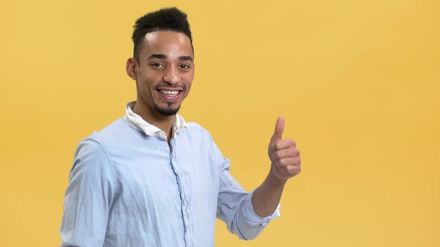 Portrait of optimistic arab guy with beard and mustache showing thumb up on camera, isolated over yellow background. Concept of emotions