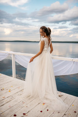 A beautiful bride is standing on the pier. Lake and cloudy sky on the background