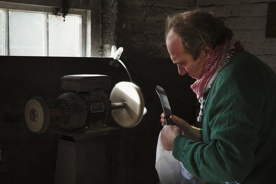 Craftsman in a workshop, holding a knife with a large blade inspecting it, and preparing to use a surface grinder.   - Powered by Adobe