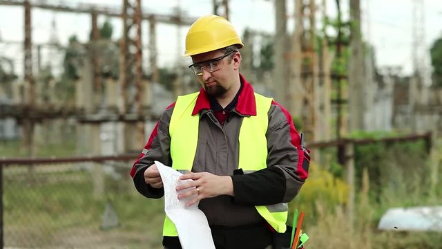 Worker with scheme of works on power station. Power man with construction documentation at power plant. Master builder in hard hat with design drawings
