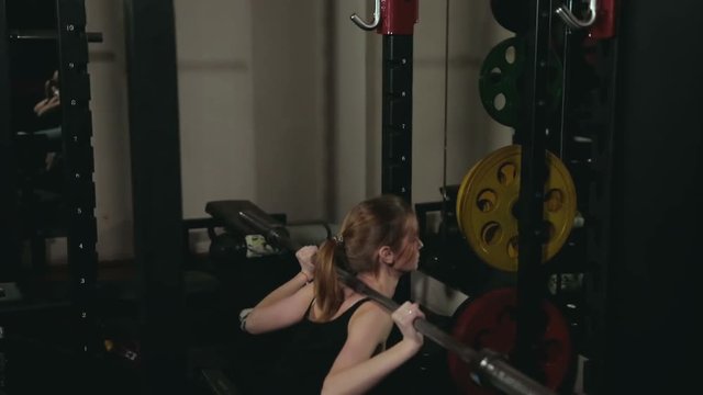 young girl with white hair in black tank top and blue tights doing squats with a vulture from a barbell in a dark fitness club