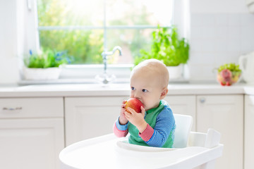 Baby boy eating apple in white kitchen at home