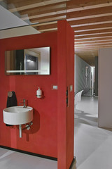 red wall behind whic there is a round wall-mounted washbasin and the mirror the floor is made of concrete and the ceiling are made of wood