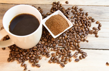 A cup of coffee with a small white ceramic dish full of ground coffee and coffee beans on wooden background.