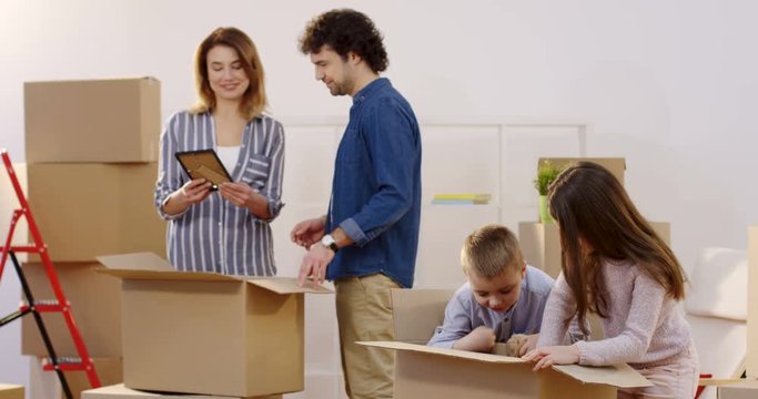 Happy young parents with their kids talking while unpacking boxes with home stuff on a moving in day. In the new flat. Indoors