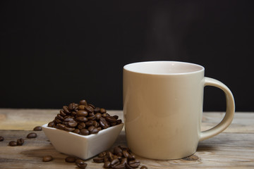 A cup of coffee with small white ceramic dish full of coffee beans on wooden bakcground.