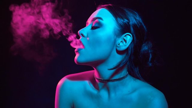 Slow motion portrait of glamour seductive gorgeous brunette woman smoking electronic cigarette in neon color light in studio