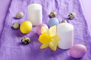Easter still life, candle, chicken and quail eggs, narcissus, on a violet textile background.