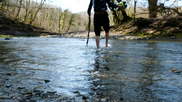 A young man crosses a mountain river wade barefoot with a stick, 4k.