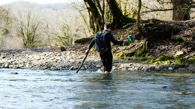 A young man with a backpack crosses a mountain river wade barefoot with a stick, 4k.