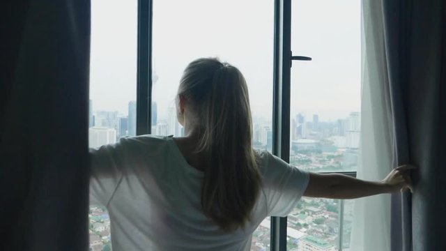 Young woman uncover the big window and looking out her apartment on the city buildings. Young girl unveil curtains in room in the cloudy morning in slow motion
