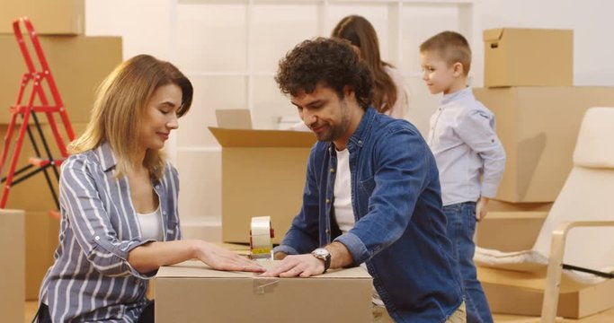 Portrait shot of the married couple packing a box with a sticky tape and their children coming to them from the background to pose together in front of the camera. Indoors
