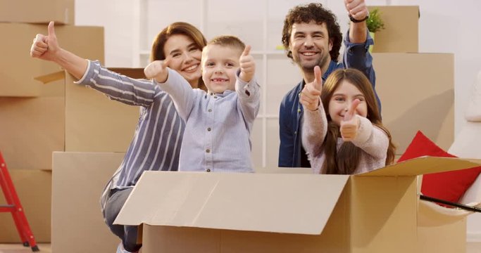 Happy good looking parents sitting together with children among boxes, smiling and showing their thumbs up. Kids sitting in the box. Moving in day. Inside