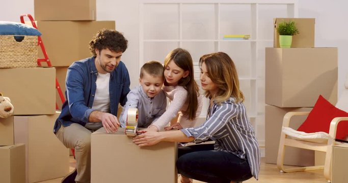 Portrait shot of the happy family of parents and two kids packing a box with a sticky tape while moving out the apartment. Inside