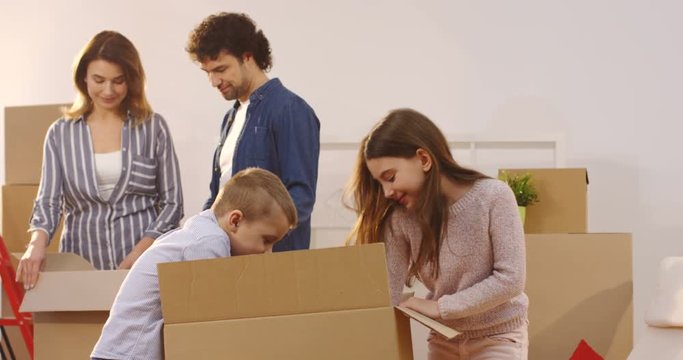 Cute sister and brother unpacking a box as they moving in the new apartment while their mother and father doing the same on the background. Indoors